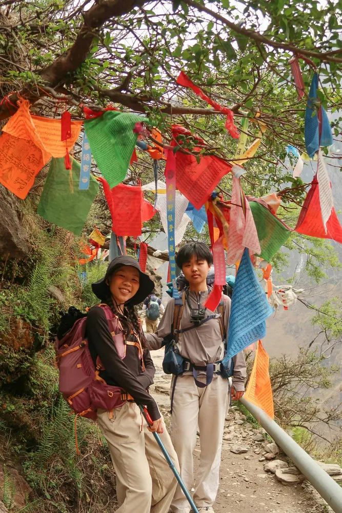 Backpackers walking along a dusty mountain path under colourful prayer flags tied to trees, posing in warm daylight