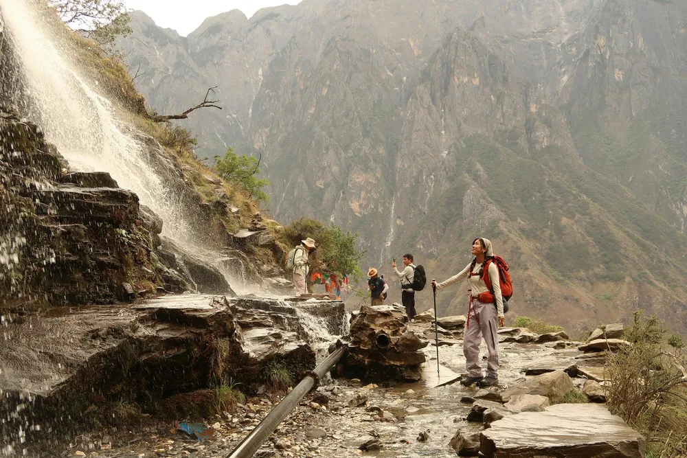 Hikers with backpacks walking along a rocky stream bed beside a wide waterfall in a steep mountain gorge