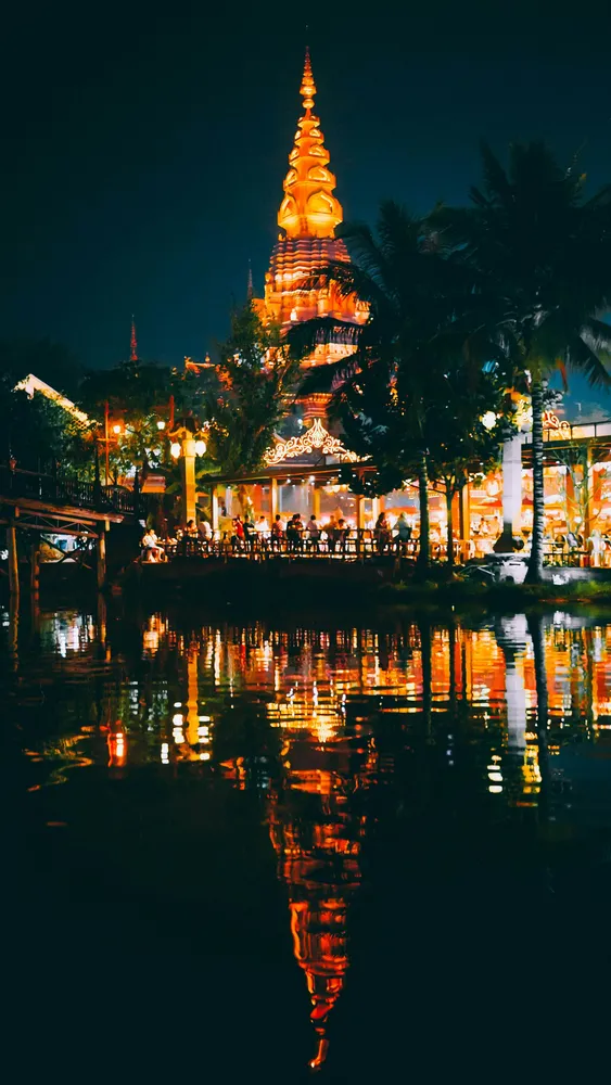 Illuminated multi-tier temple pagoda by a calm river at night, surrounded by palm trees, with golden reflections in the water