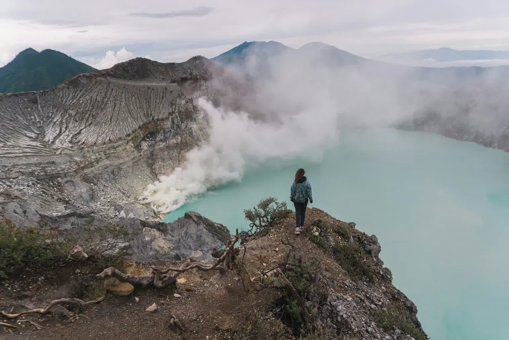 Hiker standing on a cliff edge above the turquoise sulphur crater at Banyuwangi, watching misty vapours, Mt Ijen