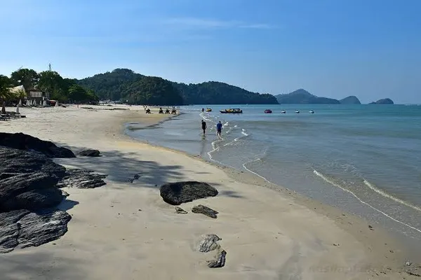 Wide sandy beach in Langkawi at low tide with scattered rocks, swimmers and walkers by the shoreline on a sunset cruise day