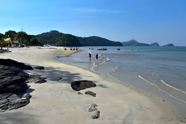 Langkawi sandy coastline with rocky foreground, gentle waves and distant islands under a bright blue sky