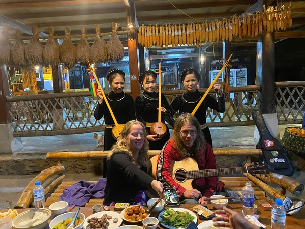 Travellers at a homestay dinner in Ha Giang, smiling around a table as an acoustic guitar plays during the Ha Giang loop