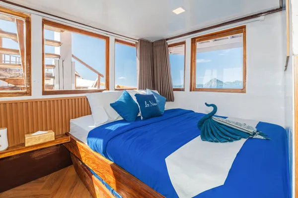 Sunlit cabin on a Komodo boat trip in Labuan Bajo, with large windows, blue bedding, and a towel swan on the bed