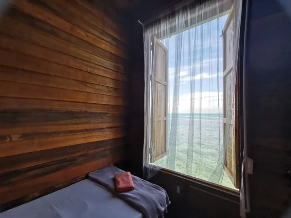 Cozy sea lodge bedroom in Semporna with a window opening to the ocean, wooden walls, curtains and a made bed