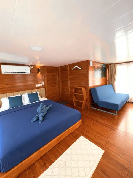 Wooden phinisi bedroom with blue bedding, wardrobe shelves and an ocean seating corner on a 3D2N cruise from Labuan Bajo, Komodo Island