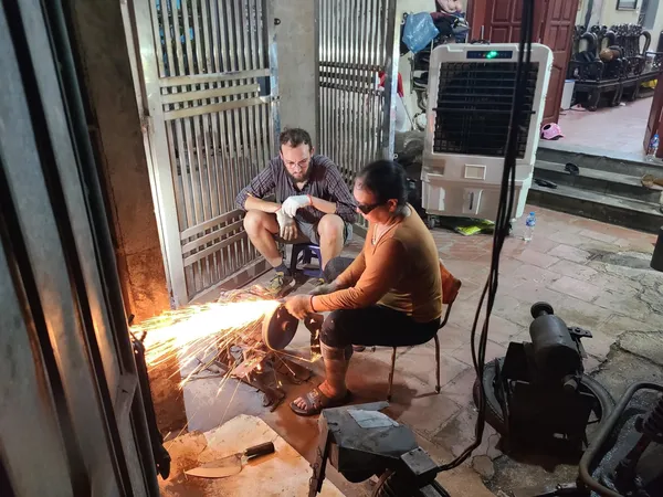 Visitor seated at the forge in Hanoi, using tools with another participant to shape their own knife amid hot metal