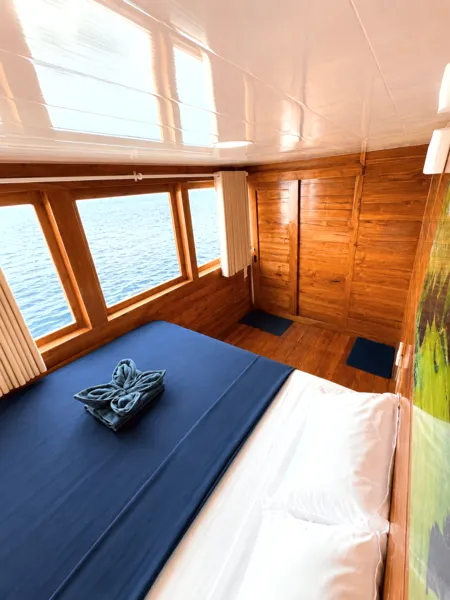 Blue-topped bunk in a wooden phinisi cabin with sea-view windows on a 3D2N shared Komodo cruise, Labuan Bajo