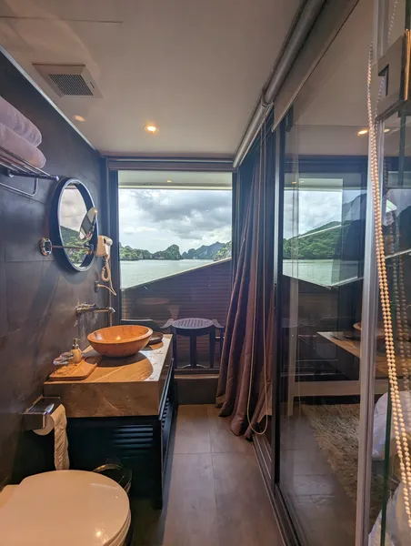 On-board en-suite bathroom with stone sink and round mirror, shower glass and karst views through the window on Halong Bay