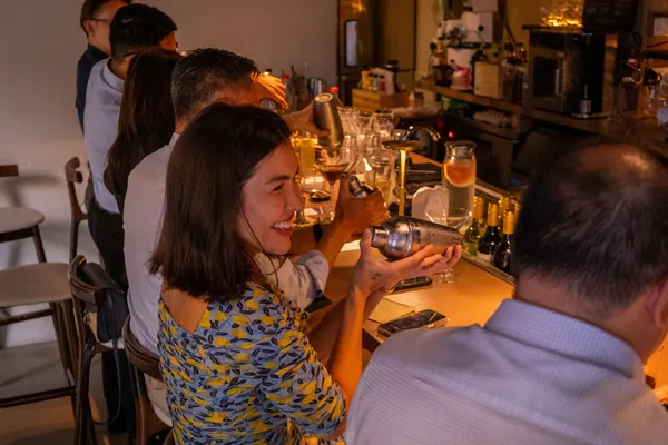 Tamu di bar bercahaya hangat di Singapura menyaksikan seorang mixologist menyajikan koktail, sambil tersenyum saat minuman dituangkan untuk private dining di Singapura