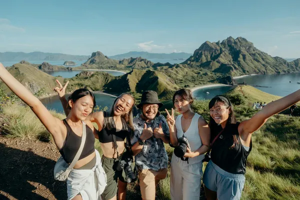 Group of travellers posing on a rugged mountain ridge with arms out, overlooking a coastal bay and distant islands