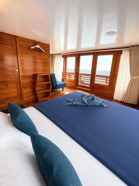 Wooden phinisi bedroom with blue-and-white beds, folded towel, and balcony doors overlooking the sea on a 3D2N cruise from Labuan Bajo, Komodo Island
