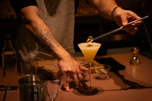 Tangan seorang mixologist menyelesaikan sentuhan akhir pada koktail di gelas coupe saat sesi cicip tiga koktail di bar intim Singapura untuk private dining.