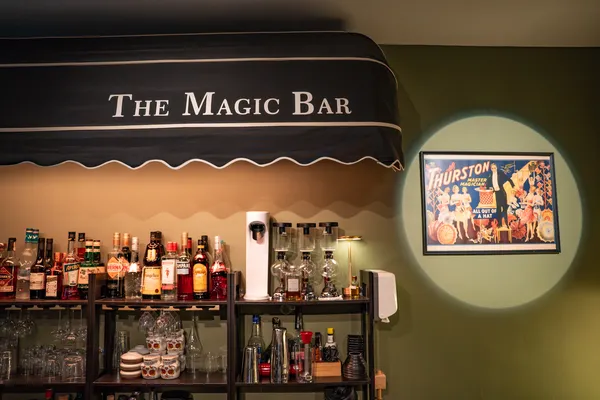 Papan tanda The Magic Bar di atas rak berisi minuman keras dan gelas di dalam bar koktel dengan pencahayaan hangat di Singapura