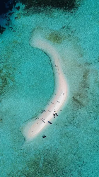 Drone perspective above a crescent beach, turquoise shallows, and moored boats lined along the sand visible from above