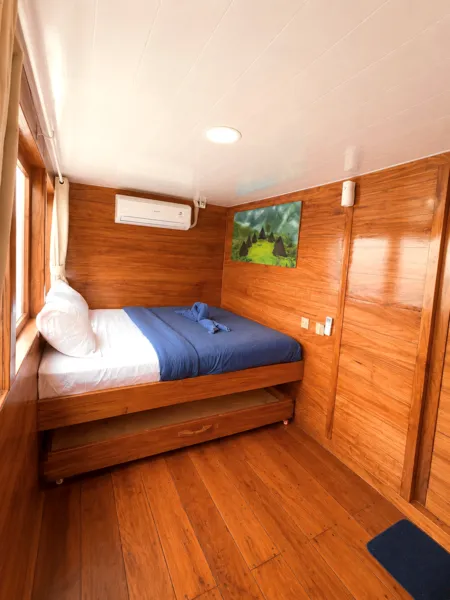 Wooden phinisi cabin with a bed, air-con unit and wall art on a 3D2N Komodo cruise, Labuan Bajo