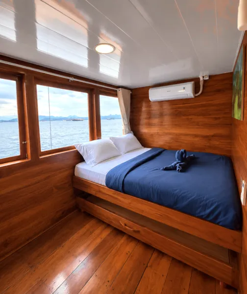 Air-conditioned phinisi cabin bedroom with blue bedding and sea-facing window on a 3D2N Komodo cruise from Labuan Bajo