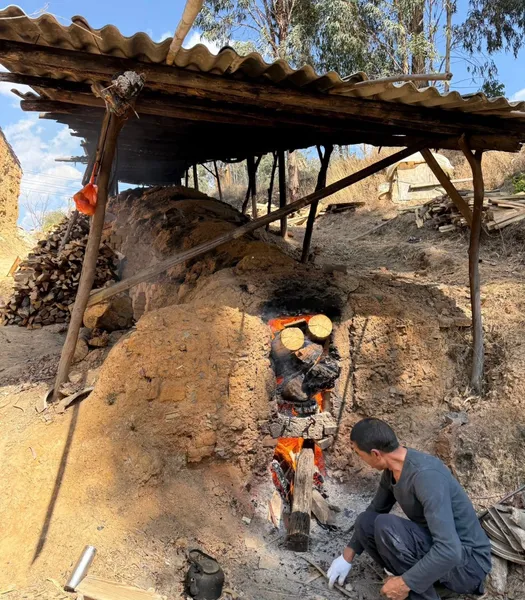 Pottery workshop outdoors in Shangri-La, as a craftsperson tends an earth kiln and stacks clay pieces under a tiled shelter