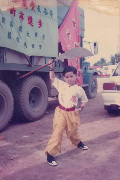 Vintage street scene: child in lion dance costume holding a wand prop beside trucks with hanging Chinese characters in Ho Chi Minh City