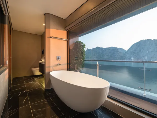 Balcony bathroom with a white freestanding soaking tub against the glass railing, with islands and mountains beyond in Halong Bay