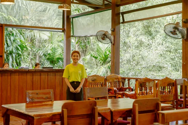Smiling woman standing by wooden dining tables inside a family-run cottage at Khao Sok Riverside Cottages, Khao Sok