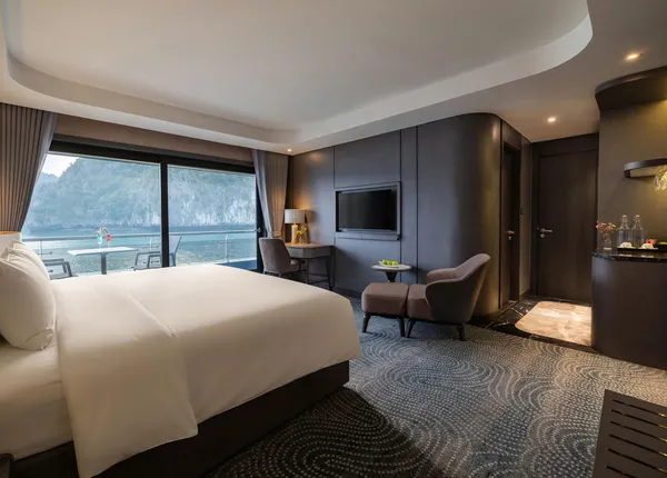 Tempat tidur kabin Tulip Cruise di dekat jendela besar, dengan area duduk untuk bersantai, menghadap Teluk Halong dalam pelayaran 2H1M