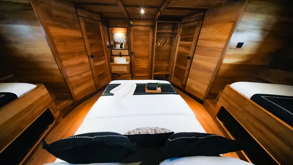 Angelica Liveaboard phinisi suite with a double bed in the foreground and an arched doorway revealing a bathroom area