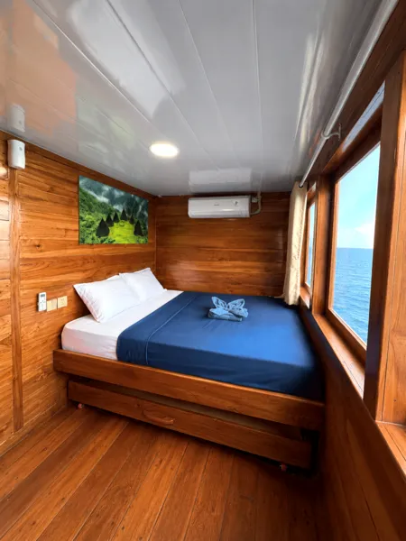 Wooden phinisi cabin bed with air con, wall art and sea-facing windows on a 3D2N shared cruise to Komodo Island