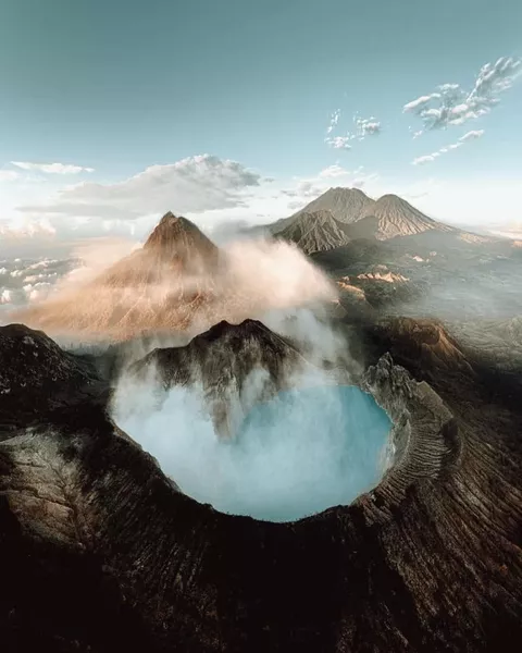 Aerial view of a steaming volcanic crater with thick smoke filling the caldera, surrounded by rugged ridges and haze