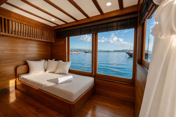 Window-side bench in a wooden phinisi cabin with anchored boats beyond the open sea views on a Rare 3D2N Komodo shared trip, Labuan Bajo