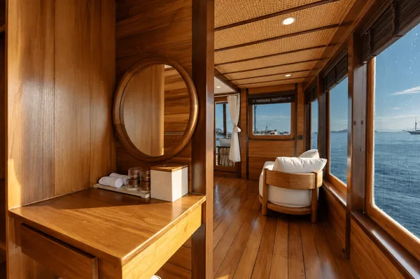 Wooden phinisi lounge corridor with oval mirror, side table and seating by the windows, with sea views on a Komodo cruise in Labuan Bajo