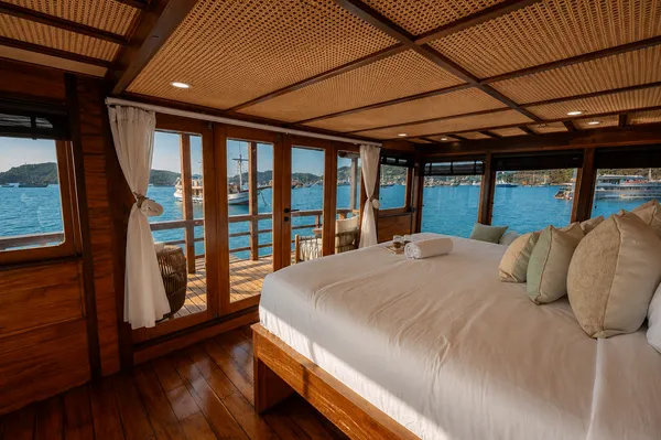 Phinisi cabin with large bed, pillows and curtained panoramic windows revealing blue water on a Komodo shared trip in Labuan Bajo