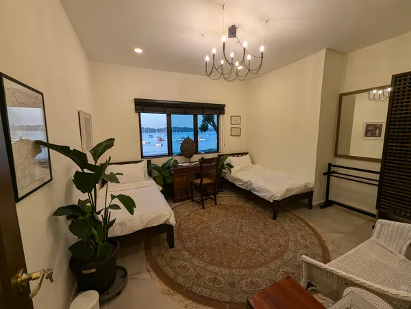 Bright bedroom with two beds, chandelier and plants, with sea views through the window at The Unexpected’s Singapore farmstay by the sea