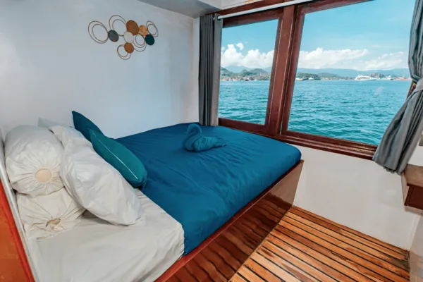 Blue-and-white phinisi cabin with a double bed by the window and ocean horizon, on a 3D2N Komodo Island adventure