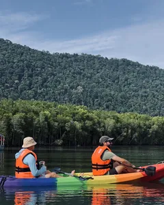 Kayakers in orange life jackets paddle along a mangrove-lined river in Kubang Badak, Langkawi, under forested hills
