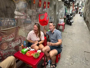Travellers seated at a low red table eating in a narrow Hanoi laneway, with dishes, drinks and street clutter