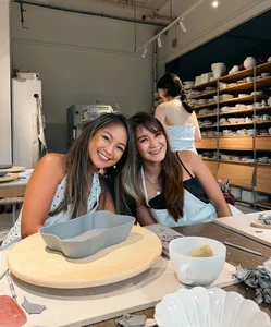 Two friends smiling at a Singapore pottery studio table with European-style ceramic plates and bowls drying behind them