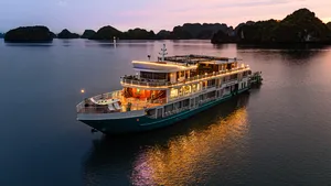 Oasis Bay party cruiser at dusk on Halong Bay with warm deck lights and silhouetted karst islands reflected in the water