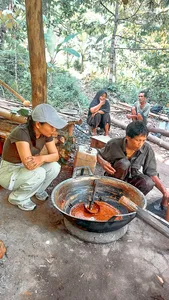Lombok forest cooking session: several people gather around a large iron bowl on a wooden platform, stirring sauce