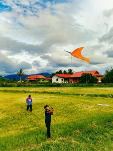 Orange kite flying over a grassy village field in Langkawi, with travellers and riverside houses under cloudy skies