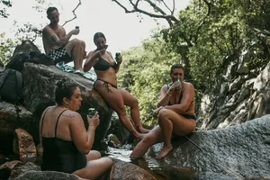 Travellers relaxing on rocks by a jungle stream, holding drinks during a Hoi An secret waterfall motorbike adventure