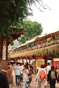 Lijiang Old Town market lane with a local guide, travellers strolling past wooden shops and rows of hanging goods under trees