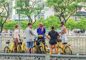 在胡志明市绿意盎然的河畔,一群骑行之旅的游客站在黄色自行车旁,与当地向导交谈