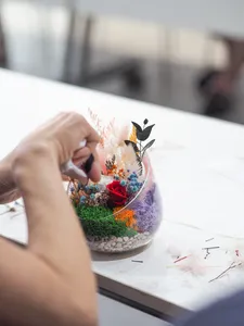 Hand placing preserved flowers into a glass floral bowl terrarium on a white tabletop during a Floral Bowl workshop in Singapore
