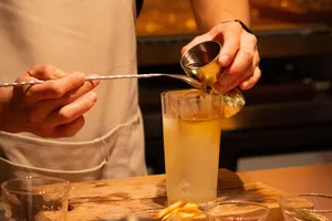 Seorang bartender menuangkan campuran berwarna amber ke dalam gelas tinggi di bar koktail Singapura, sambil memegang sendok pengaduk panjang