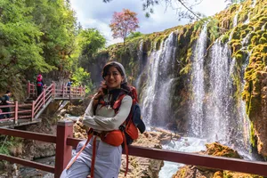 Hiker posing by a multi-tier waterfall in Shangri-La, standing on a wooden platform with red handrails and trekking gear