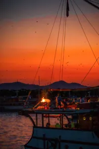 A boat deck at dusk with a small fire and fruit bats circling overhead, sails and ropes frame the sky, Komodo Island