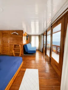 Wooden cabin interior with a blue sofa chair, bed and sea-facing windows, showing comfort for a 3D2N shared Komodo cruise, Komodo Island