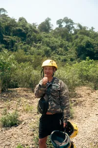 Traveller in a yellow helmet and camouflage stands on a rocky trail through Phong Nha jungle, holding up a snack