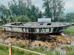 Old, damaged boat resting in shallow muddy water and grass, with trees and a sign in a quiet riverside area